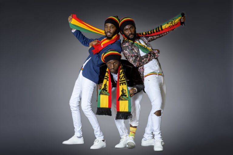Three band members in white outfits and rainbow caps posed against a gray studio backdrop