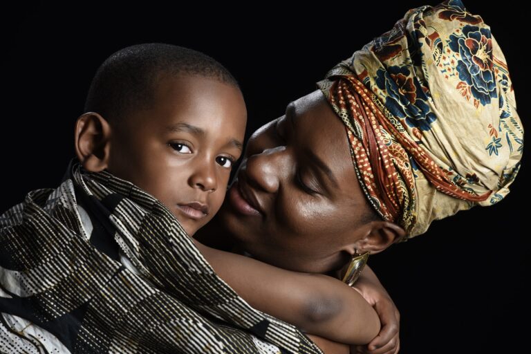 Mother holding a child, both wearing headwraps, against a black background