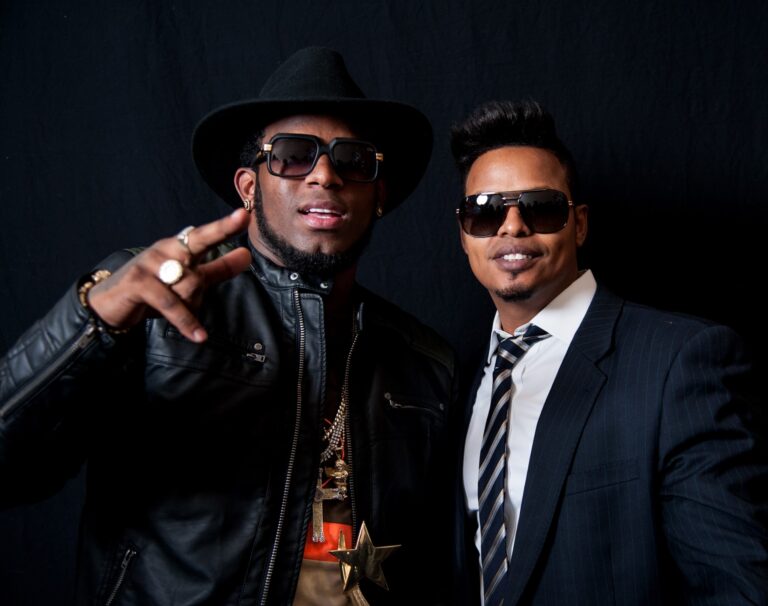 Two men in black outfits, hats, and glasses posed against a dark background