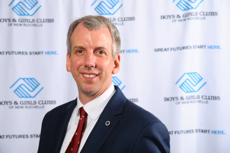 Headshot of a smiling man in a suit in front of an event backdrop