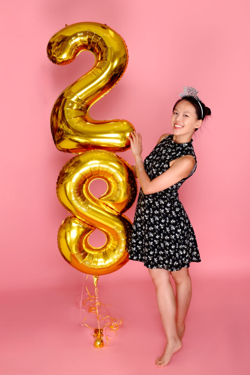 Birthday portrait holding gold 28 balloons against a pink studio backdrop