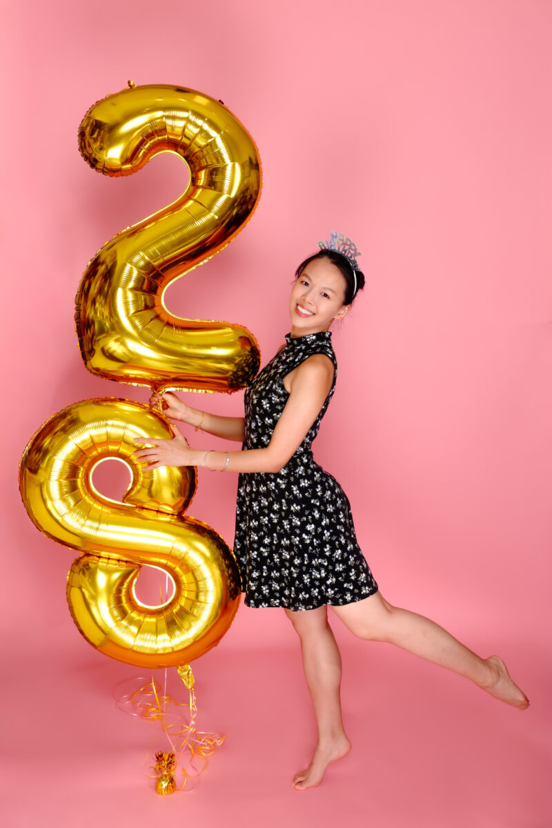 Playful birthday portrait holding gold 28 balloons with one leg raised