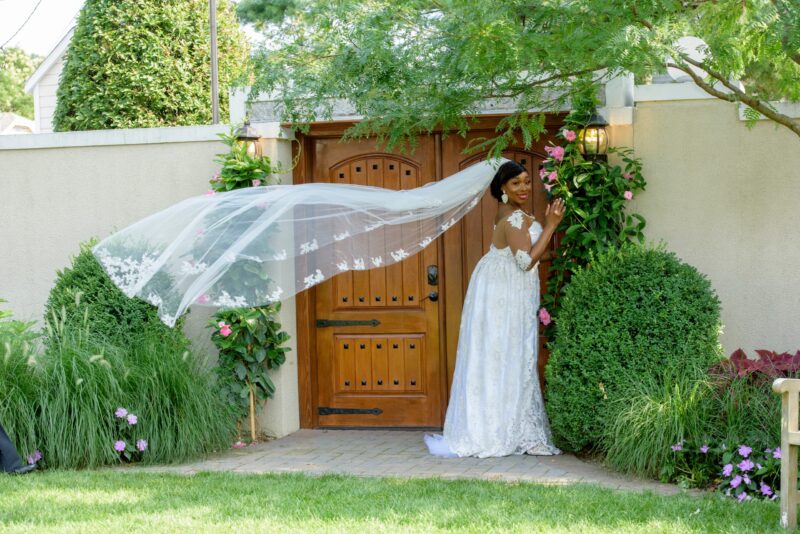 Bride standing beside a garden entrance with her veil flowing through the air