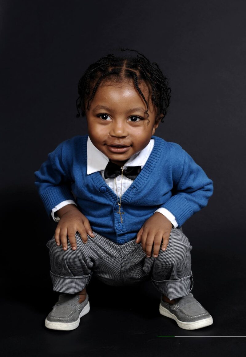 Child crouching in a blue cardigan and bow tie against a dark studio backdrop