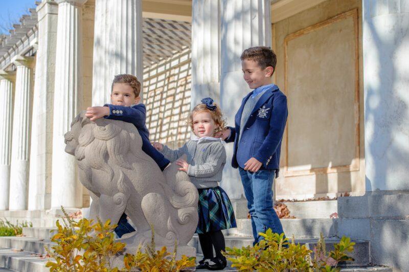 Three children in formal clothes posing beside a stone lion outside a building