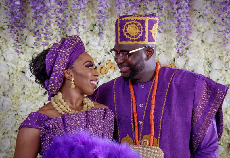 Couple wearing matching purple celebration attire in front of a floral backdrop