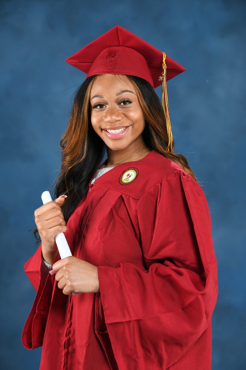 Graduate holding a diploma in a red cap and gown against a blue studio backdrop